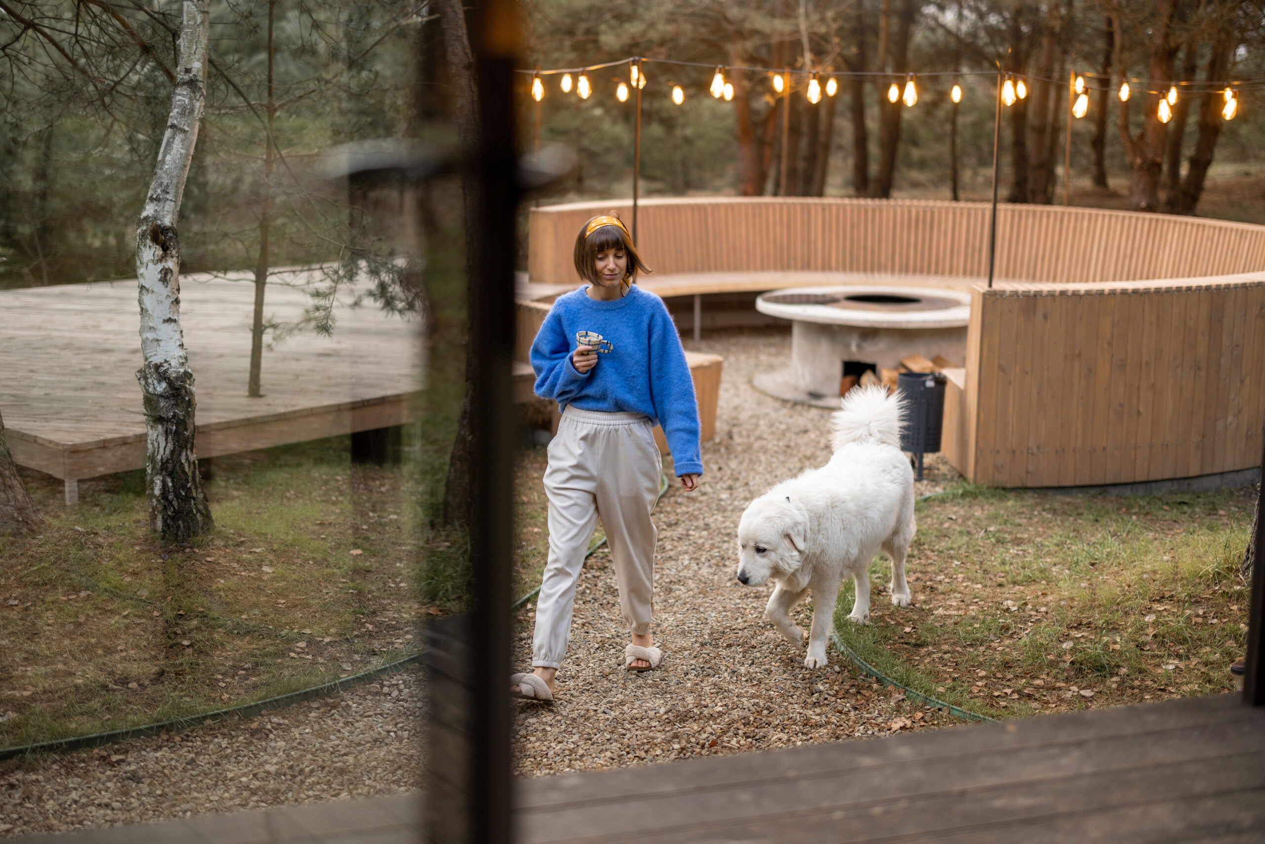 Woman walks with a dog at cozy backyard with luxury fireplace and round lounge bench in pine forest at dusk. Concept of rest and leisure time at countryside